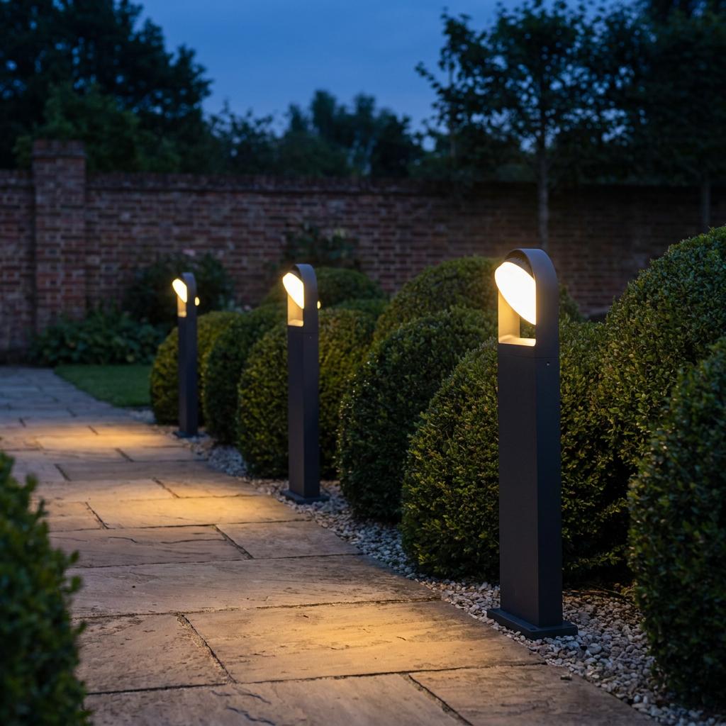 Three grey bollard lights illuminate a stone garden path lined with rounded hedges at dusk.