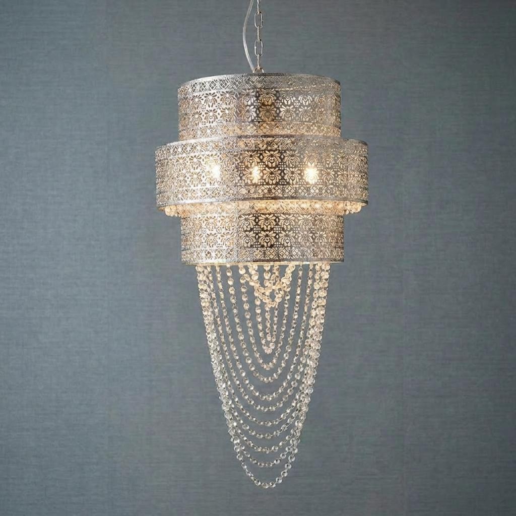 Chrome tiered pendant light with intricate cutouts and cascading crystal bead chains against a textured grey wall.