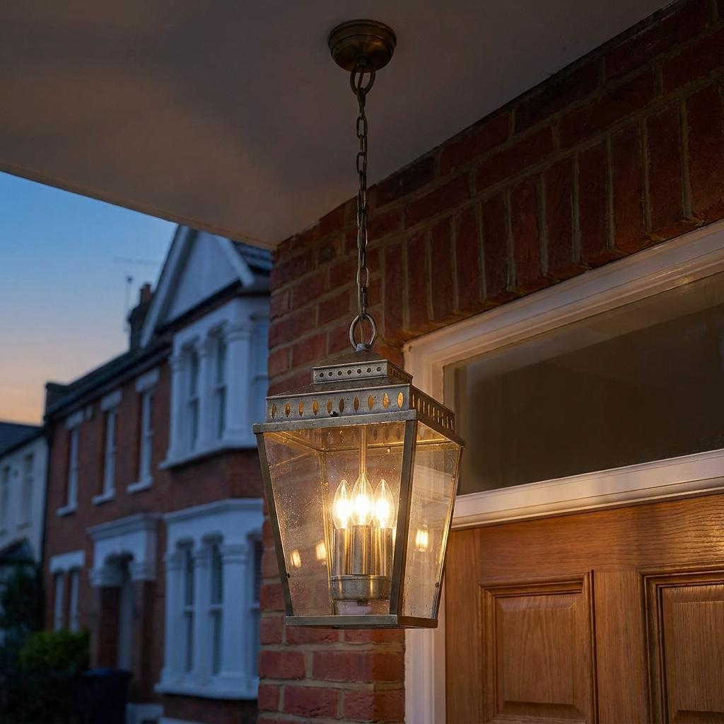Brass outdoor chain lantern with three lit candle bulbs hanging from a porch ceiling next to a brick wall.