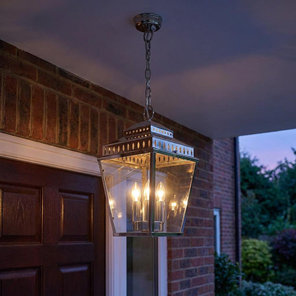 A polished nickel 3-light chain lantern hanging on an outdoor porch against a brick wall at dusk.