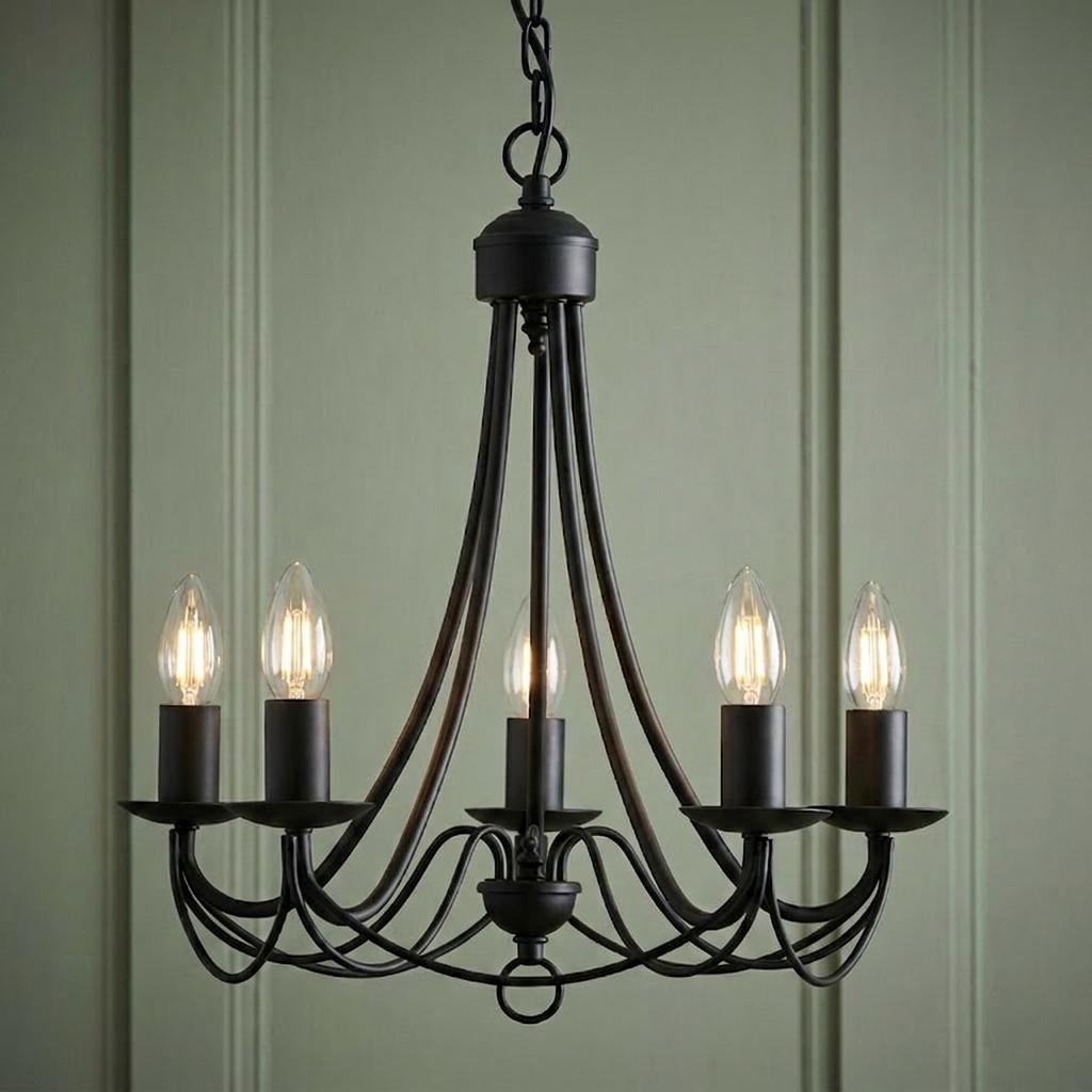 Five-light matt black wrought iron chandelier with glowing candle bulbs against a green paneled wall.