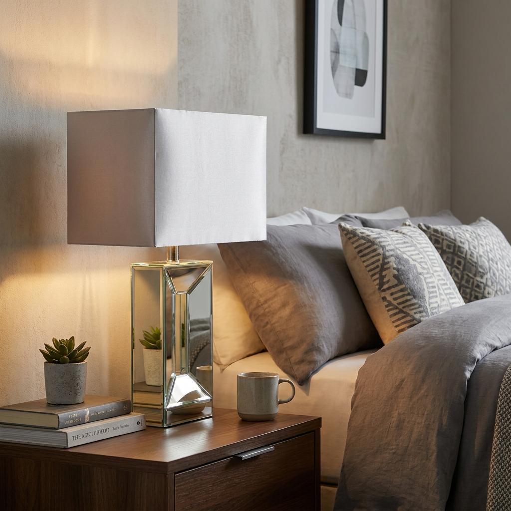 Mirrored geometric table lamp with a grey rectangular shade on a bedside table in a modern bedroom setting.