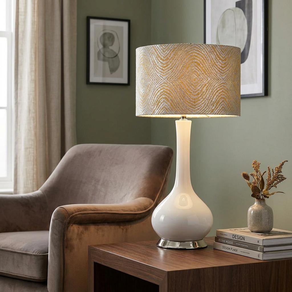 White ceramic gourd-shaped table lamp with a silver patterned shade on a wooden table next to a plush armchair.