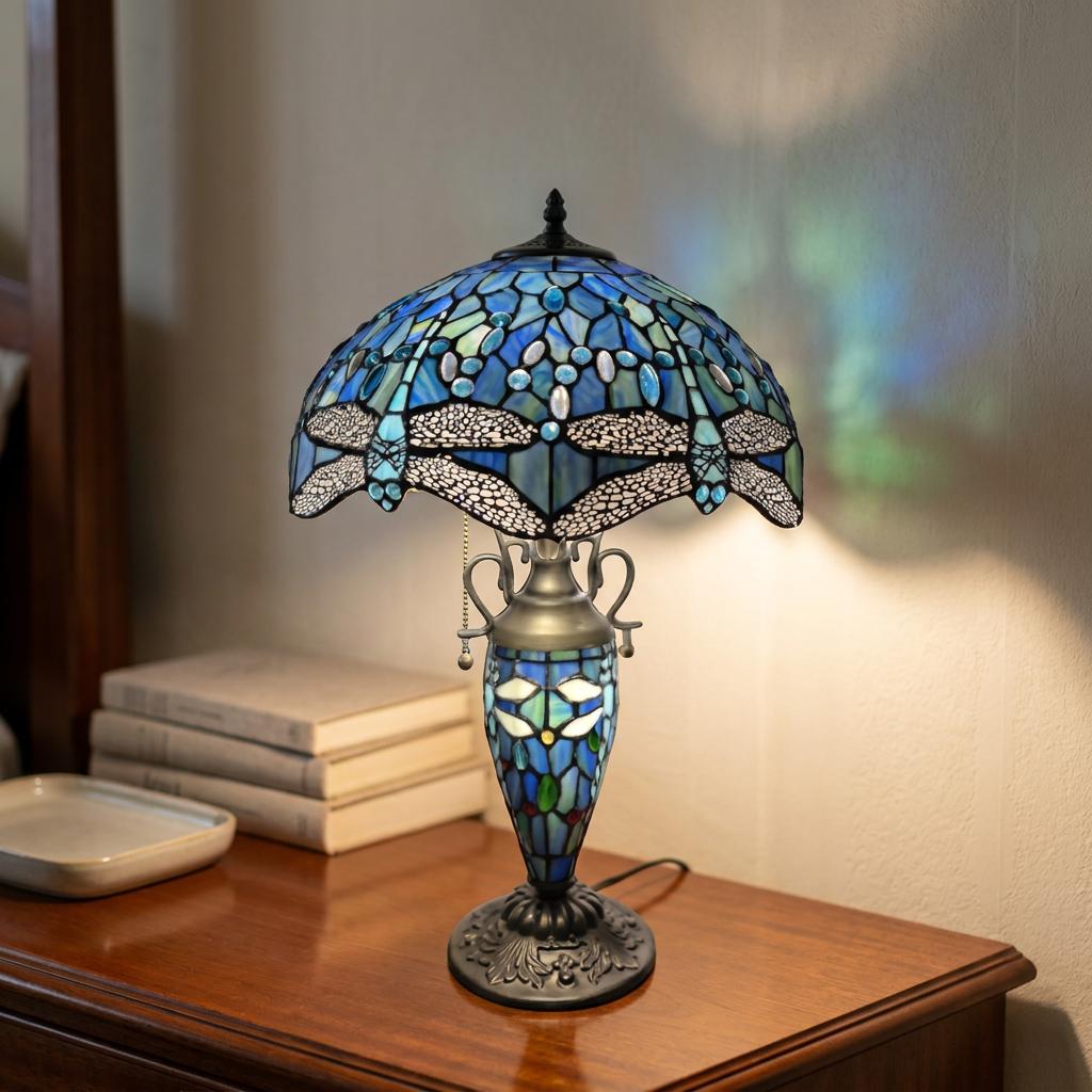 Blue dragonfly stained glass Tiffany table lamp lit on a wooden nightstand beside a stack of books.