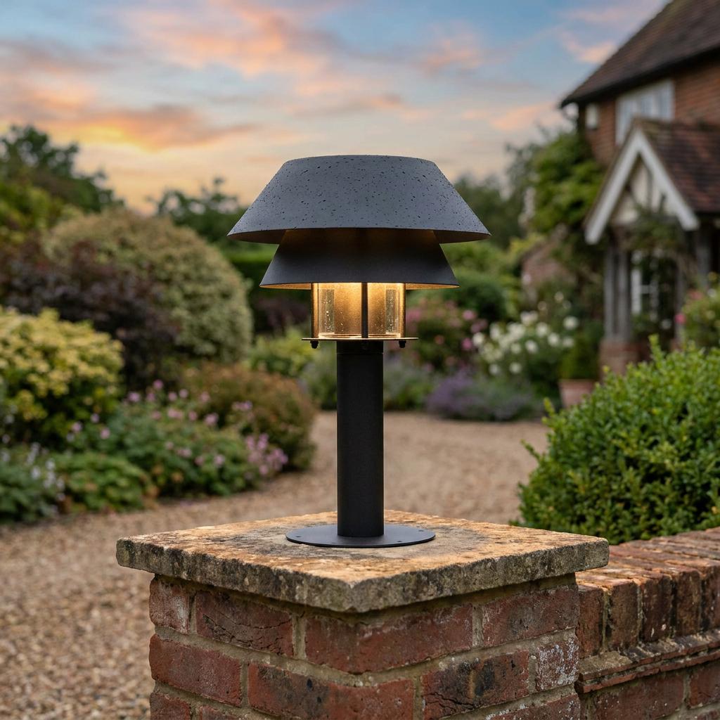 Modern black tiered pedestal light glowing on a brick wall pillar in a garden at twilight.