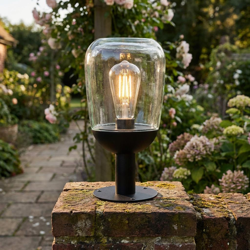 Modern outdoor pedestal light with clear glass shade and glowing filament bulb on a garden brick wall.