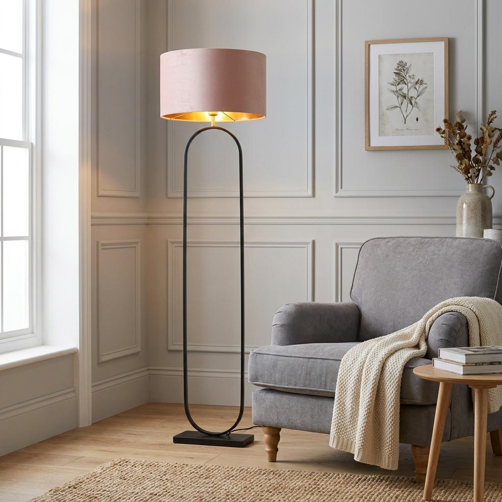 A modish matt black floor lamp with a pink velvet shade and gold interior stands next to a grey armchair.