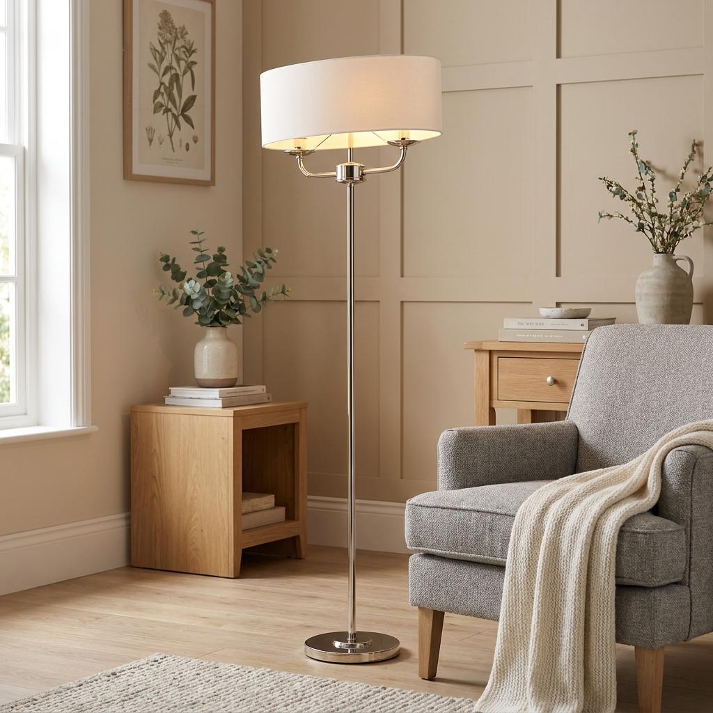 Nixon bright nickel floor lamp with a white drum shade stands in a modern living room next to a grey armchair.