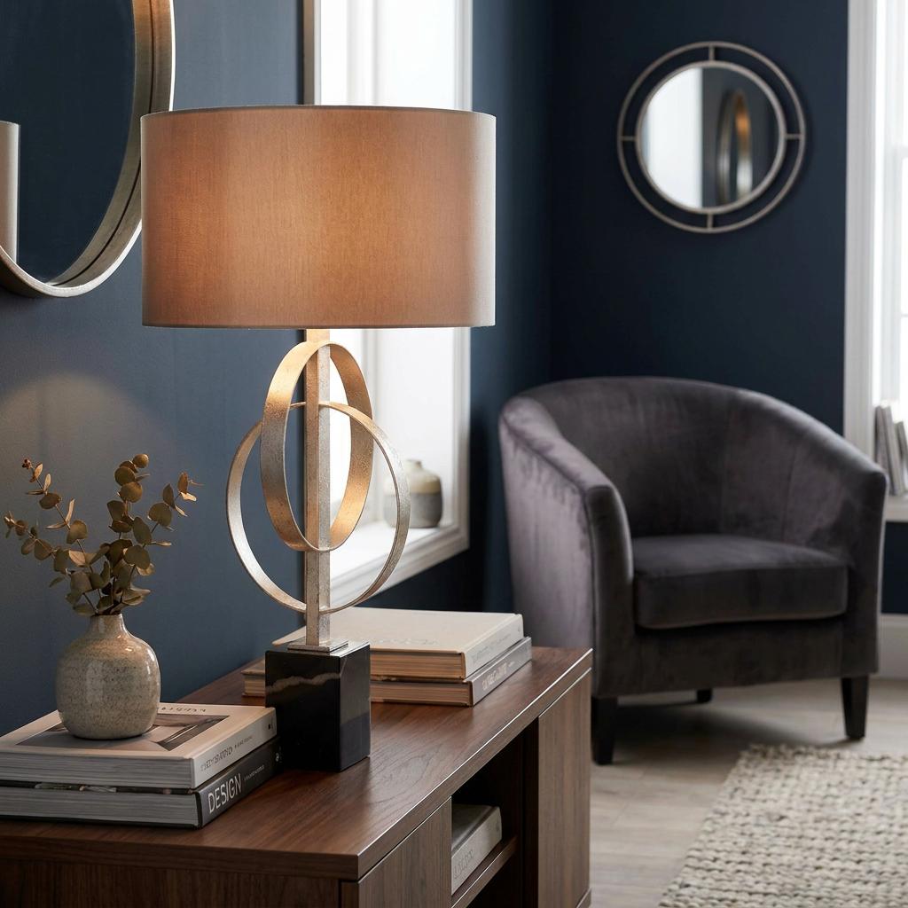 Norfolk silver table lamp with interlocking ring base and mink shade on a wooden console in a moody blue living room.
