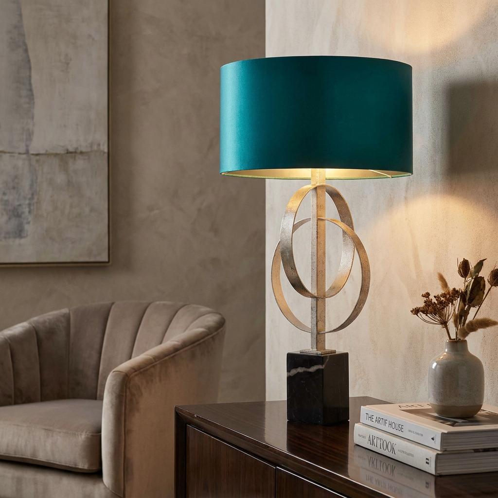 Silver interlocking ring table lamp with a teal shade and black marble base on a sideboard in a cozy room.