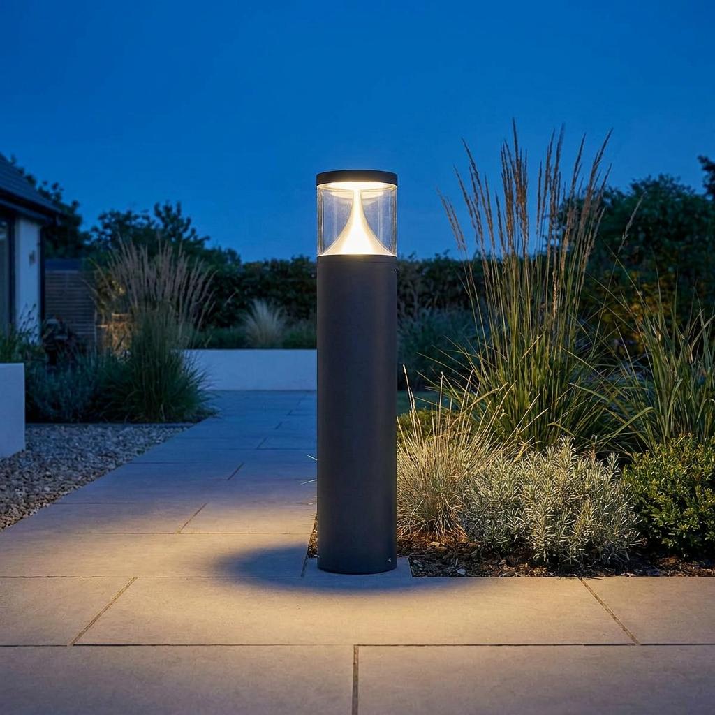 Illuminated graphite cylindrical bollard light on a stone pathway in a garden at dusk.