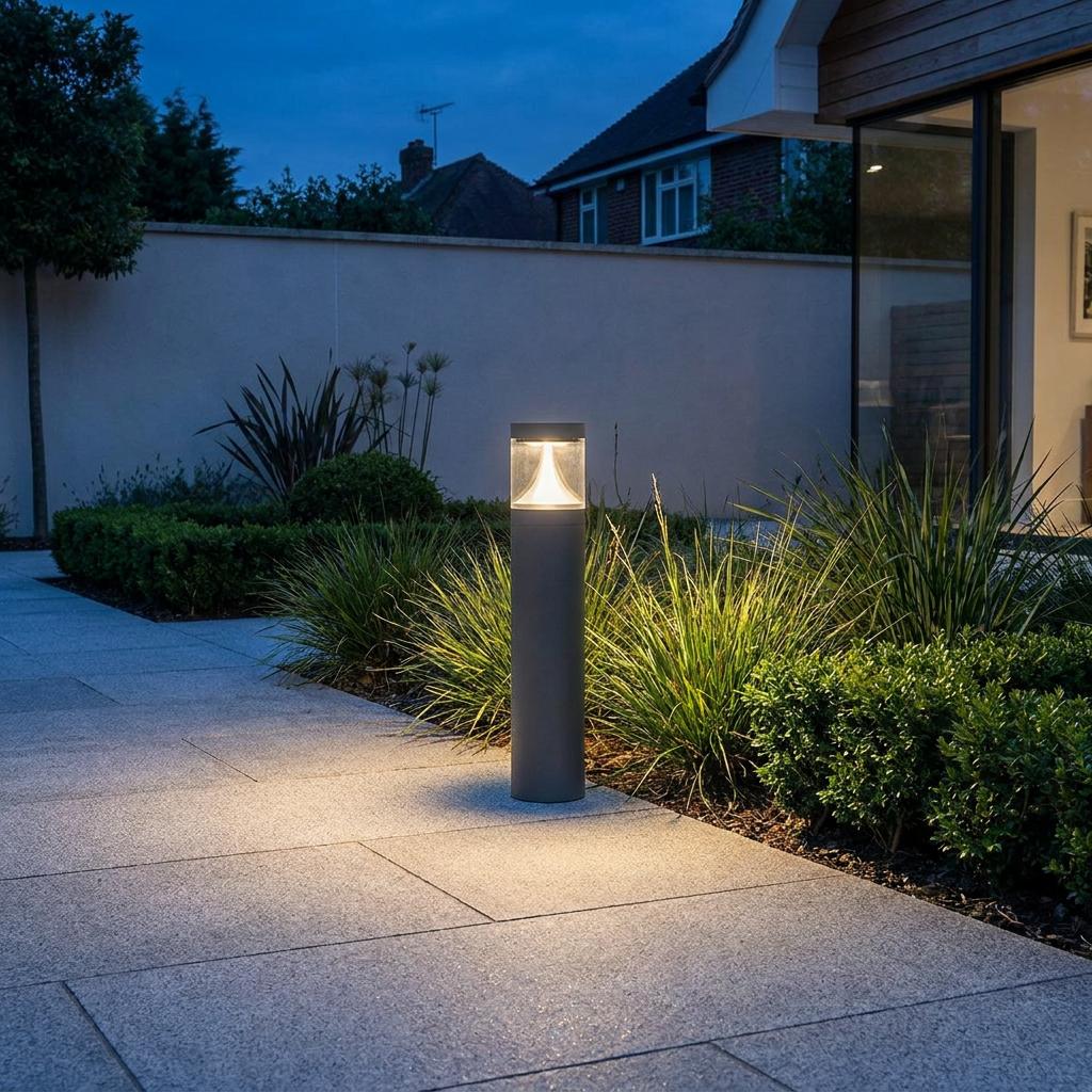 A graphite bollard light illuminates a stone path and garden greenery with a warm glow at dusk.