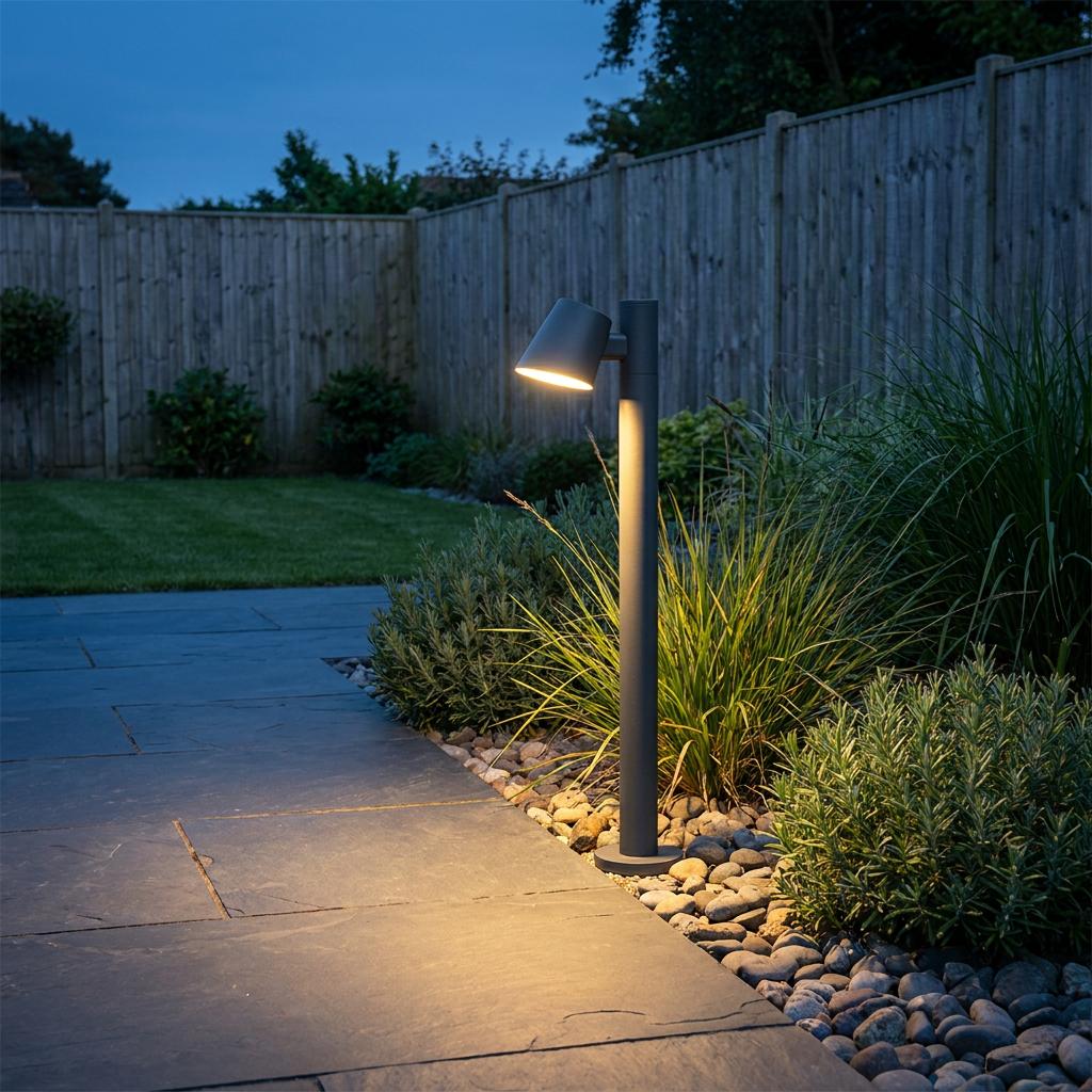 A graphite outdoor bollard light illuminating a slate patio and garden plants at dusk.