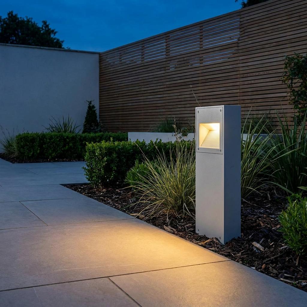 Modern grey bollard light casting a warm glow onto a stone path in a landscaped garden at dusk.