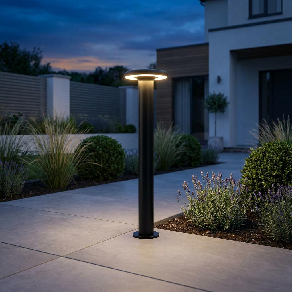 Black LED bollard post with a round glowing head illuminates a stone path in a modern garden at twilight.