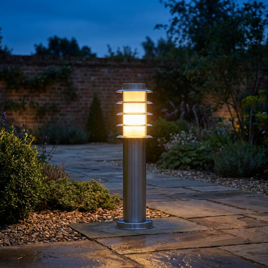 A stainless steel outdoor bollard light with tiered louvers glows on a stone garden path at dusk.