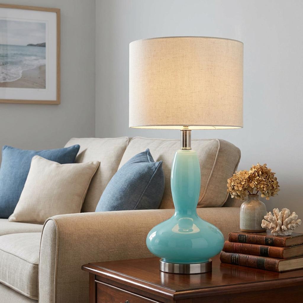 Sea blue glass touch table lamp with a beige fabric drum shade on a wooden side table in a cozy living room.