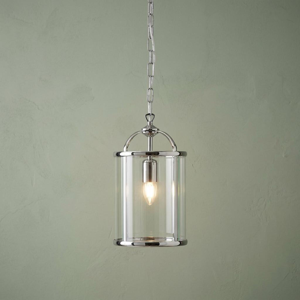 Chrome cylindrical lantern pendant light with a clear glass shade and single glowing bulb against a green wall.