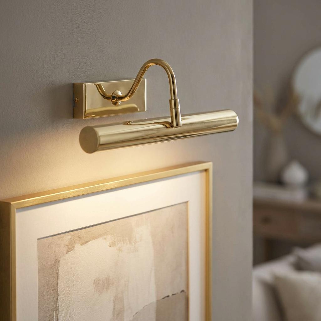 Polished brass LED picture light mounted on a wall, illuminating a framed artwork in a modern interior.