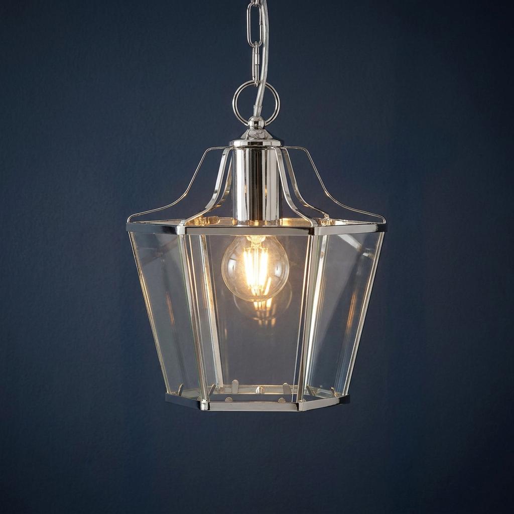 A polished chrome and clear glass lantern ceiling pendant with a glowing bulb against a dark blue background.