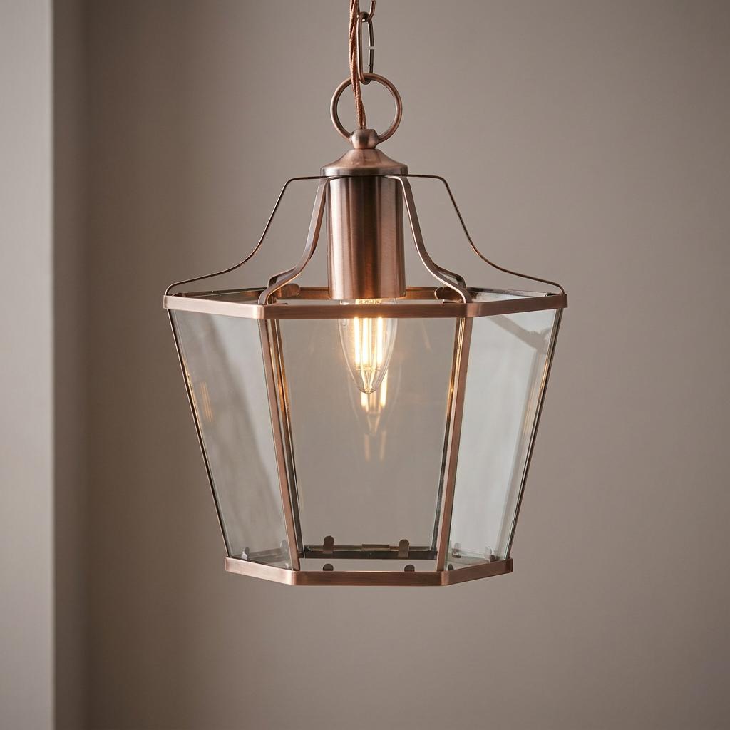 Copper and clear glass hexagonal lantern pendant light with a lit Edison bulb hanging against a grey wall.