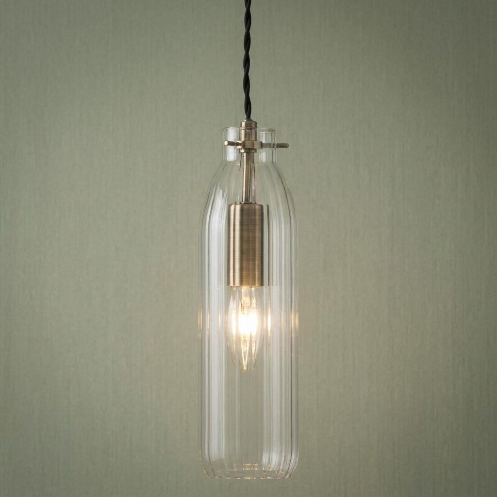 Antique brass pendant light with a fluted clear glass bottle-shaped shade against a green wall.