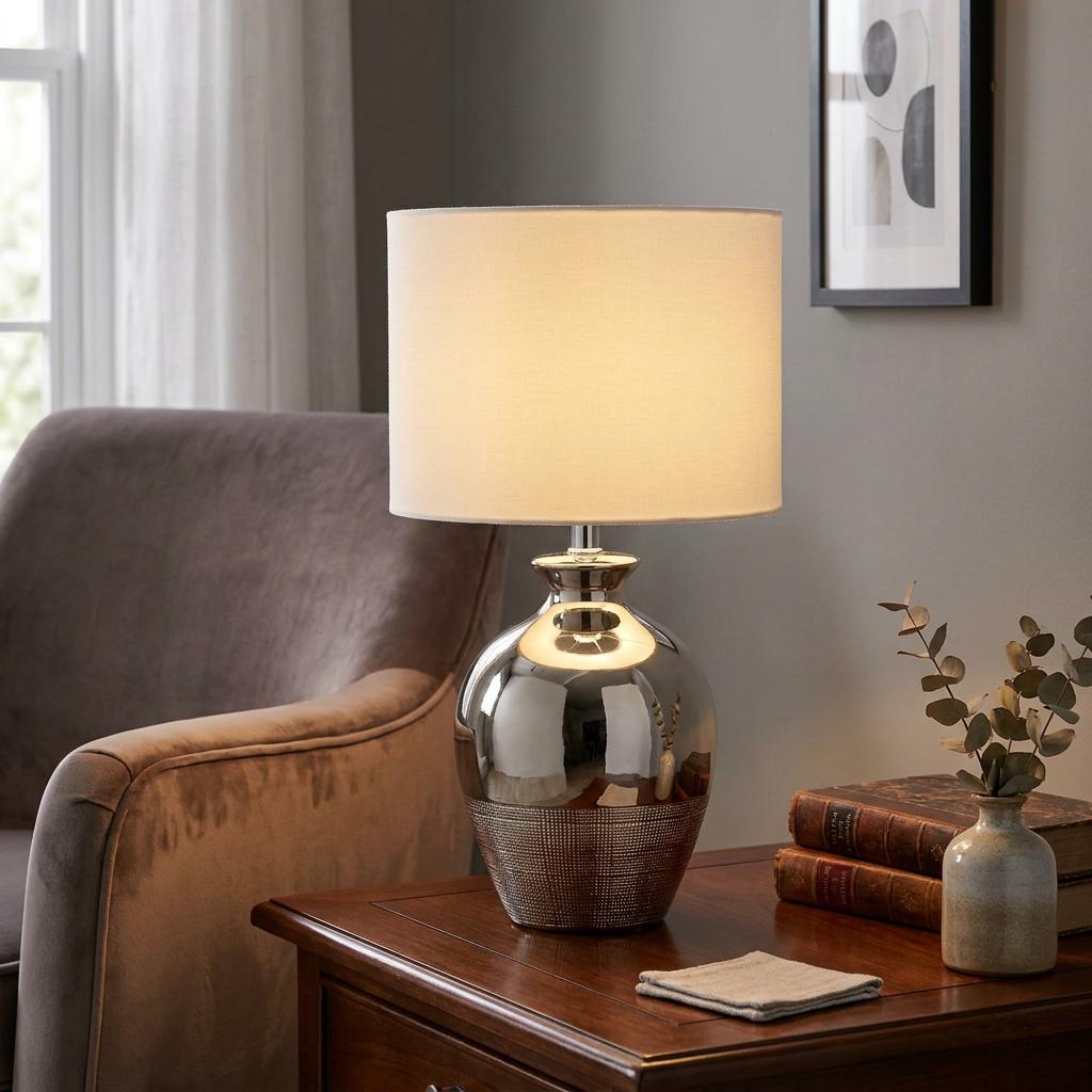 Metallic ceramic chrome table lamp with a cream drum shade glowing on a wooden table next to an armchair.