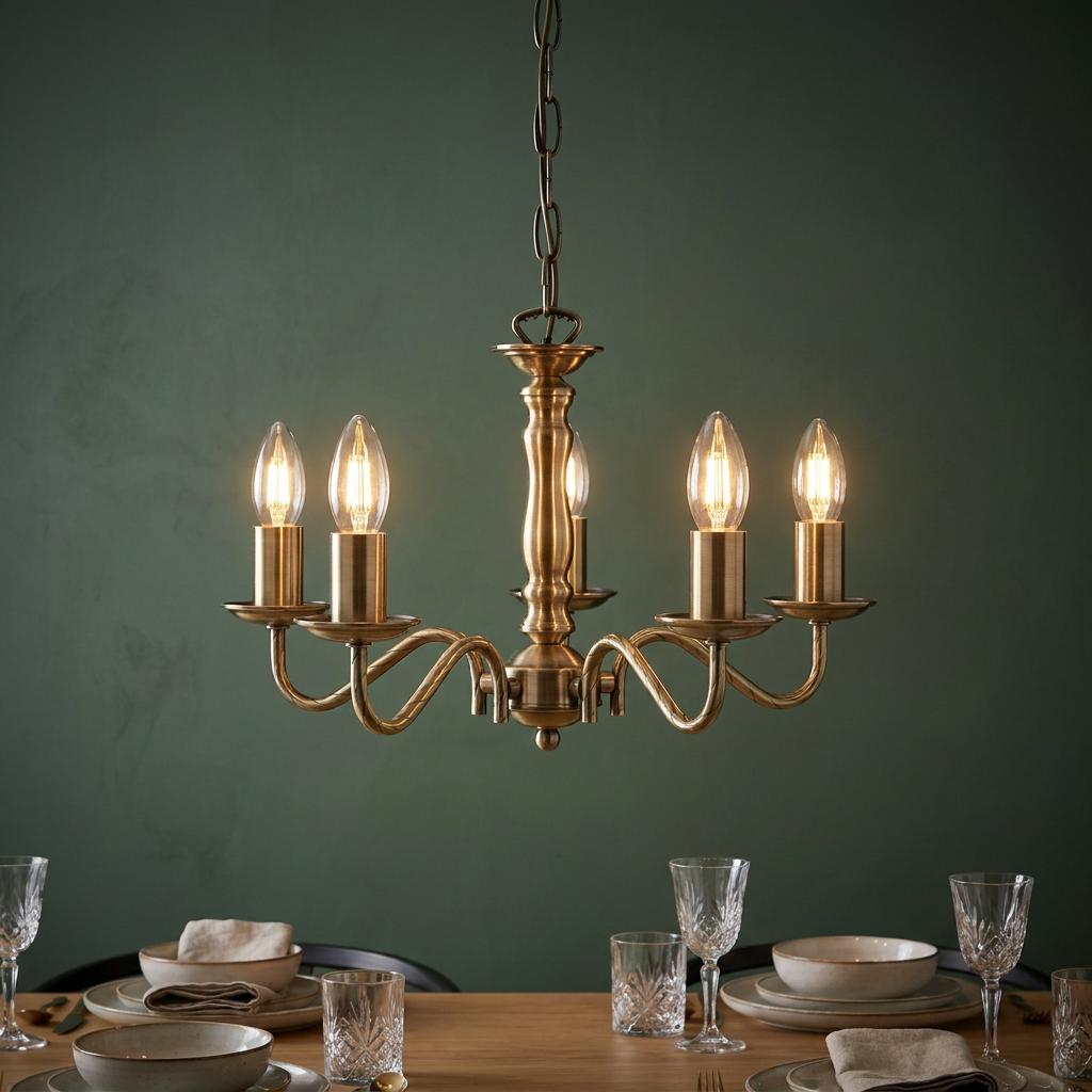 Five-light antique brass chandelier with glowing candle bulbs hanging above a set dining table with a green wall.