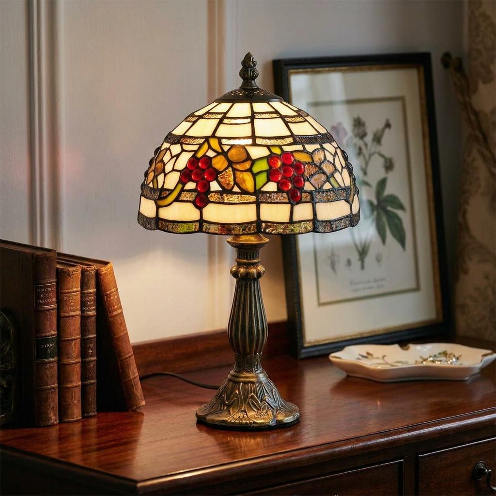 Stained glass Tiffany lamp with a red grape motif on a wooden desk next to vintage books and a framed picture.