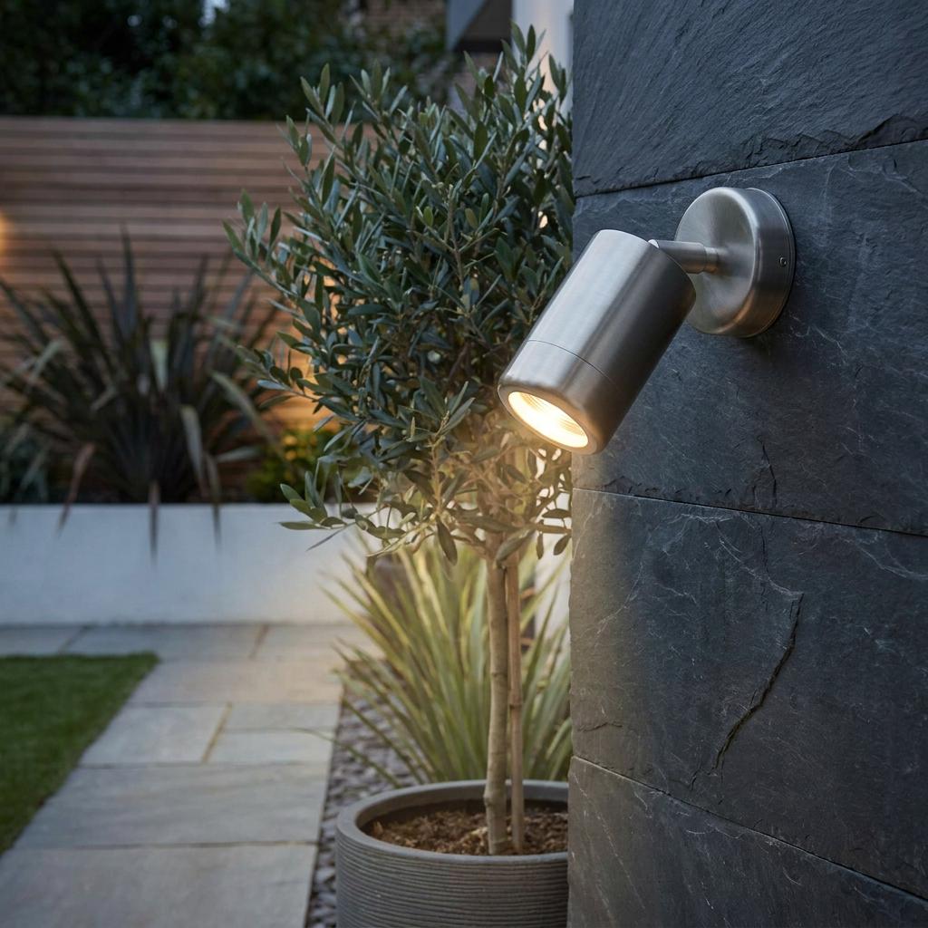 Stainless steel outdoor spotlight mounted on a slate wall, illuminating an olive tree in a modern garden.