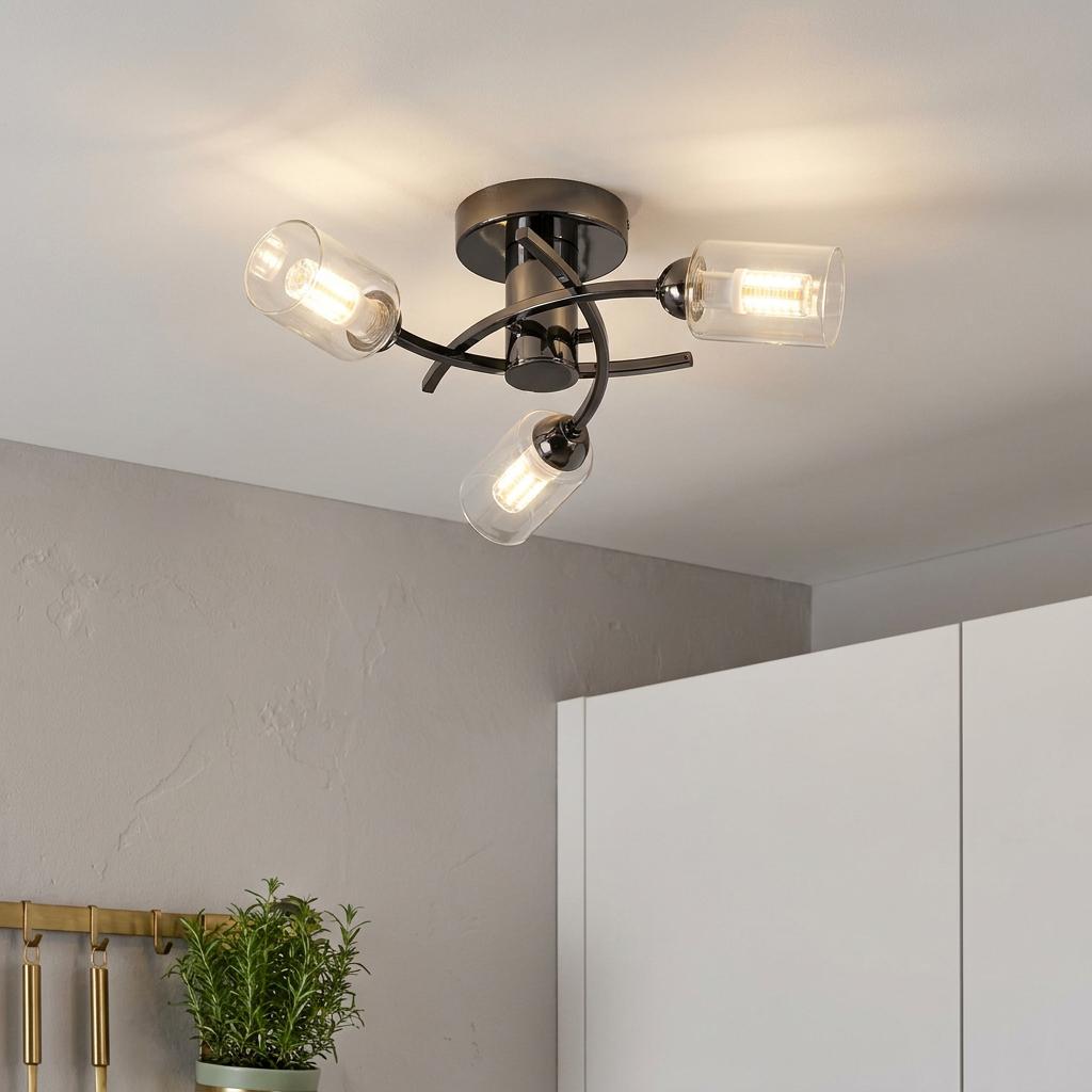 A black three-light flush ceiling fixture with curved arms and clear cylindrical glass shades lit in a kitchen.