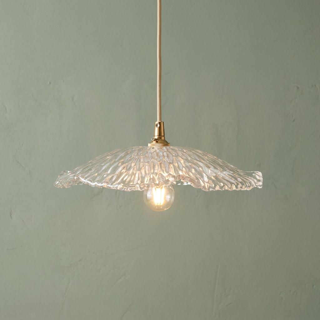Clear textured glass orchid pendant shade with brass hardware and a lit bulb against a sage green wall.