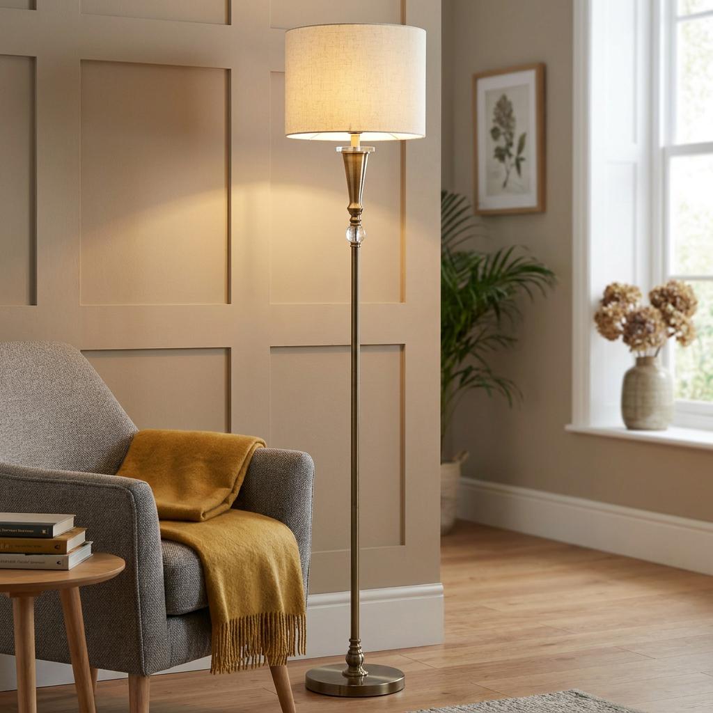 An antique brass floor lamp with a cream fabric drum shade stands in a cozy living room next to a grey armchair.