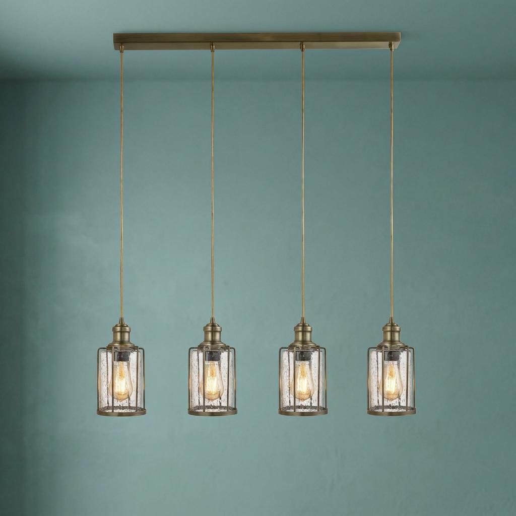 Four industrial brass pendant lights with seeded glass shades and wire cages against a muted teal wall.