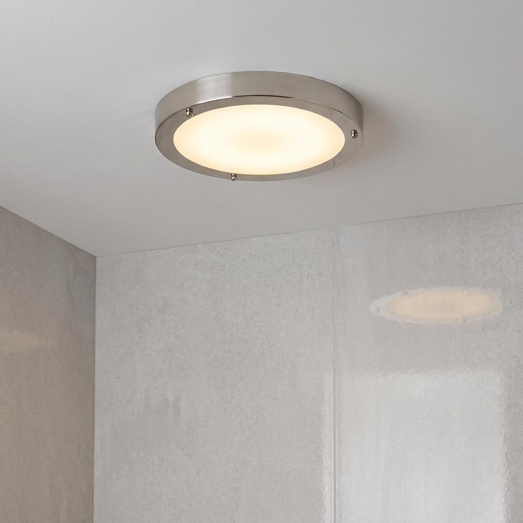 A circular brushed nickel and frosted glass flush-mount ceiling light illuminated on a white bathroom ceiling.