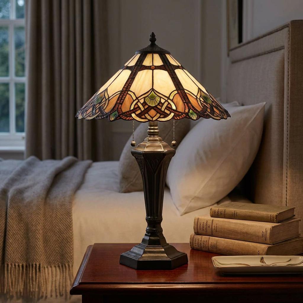 Tiffany-style table lamp with an ornate stained glass shade glowing on a wooden bedside table next to a bed.