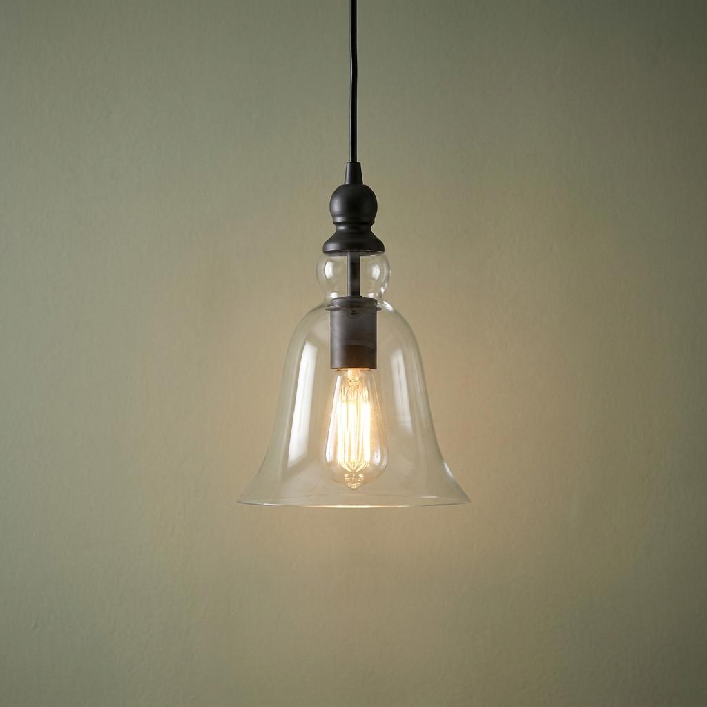 Black pendant light with a clear bell-shaped glass shade and a glowing Edison bulb against a sage green wall.