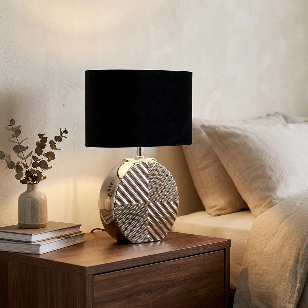 Regent chrome table lamp with a textured geometric base and black shade on a wooden nightstand by a bed.