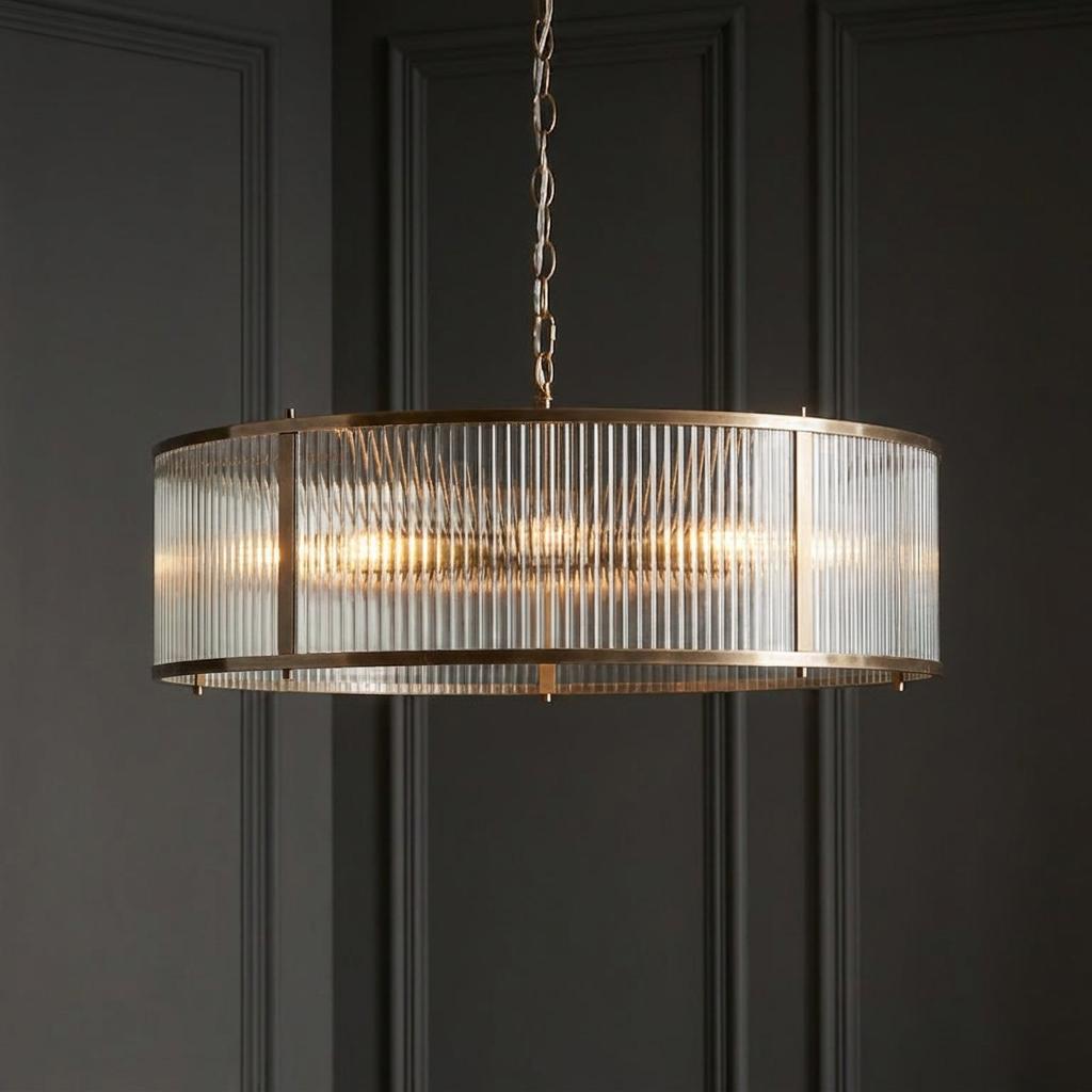 Ridgeton 8-light antique brass pendant with a circular ribbed glass shade, glowing against a dark paneled wall.