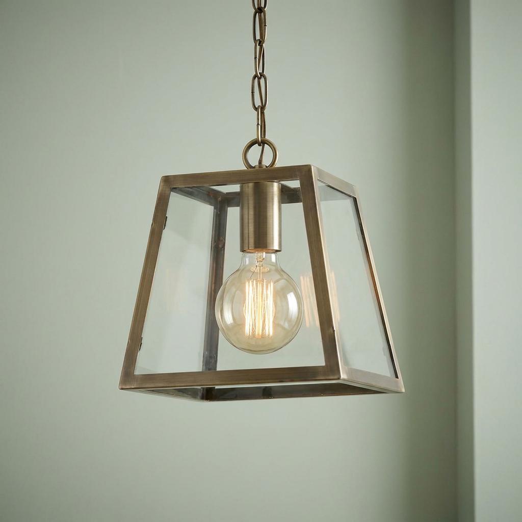 Antique brass lantern-style pendant light with glass panels and a glowing filament bulb against a pale green wall.