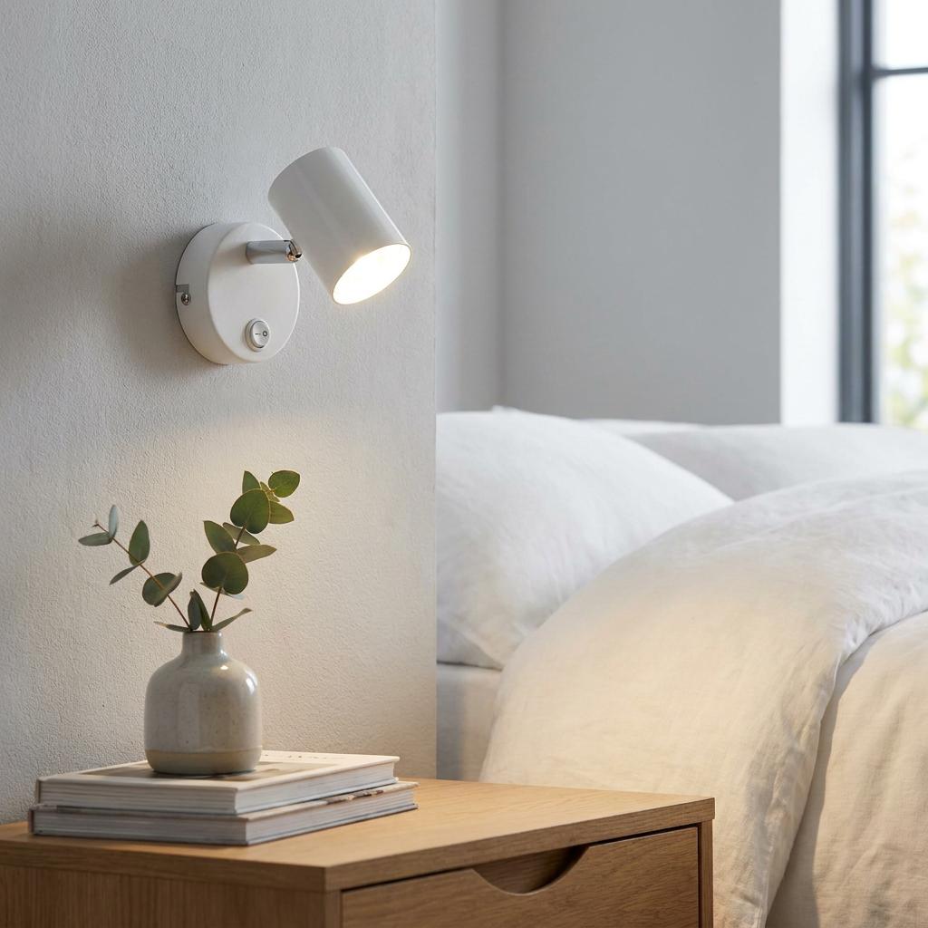 White cylinder adjustable wall spotlight illuminating a bedside table with a plant and books.