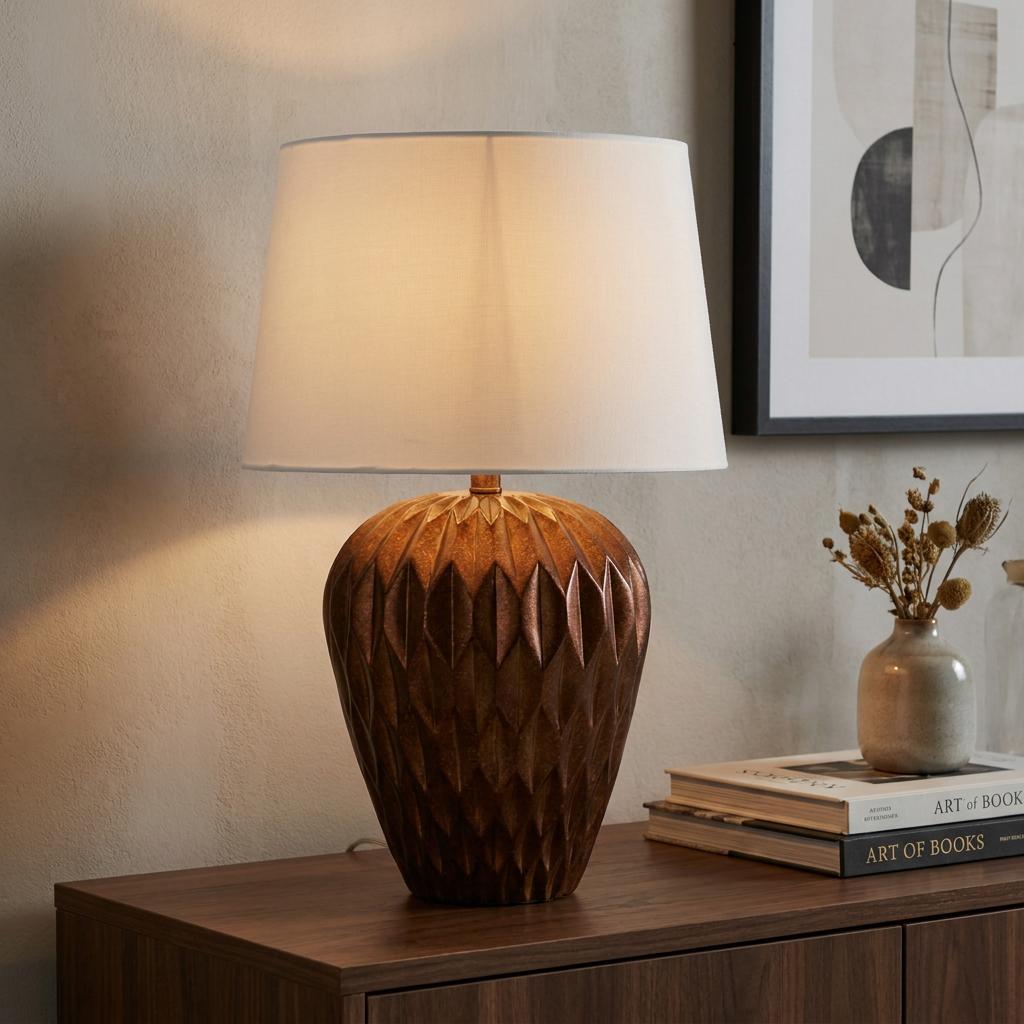 Table lamp with a textured bronze resin base and white drum shade, set on a wooden sideboard with books.