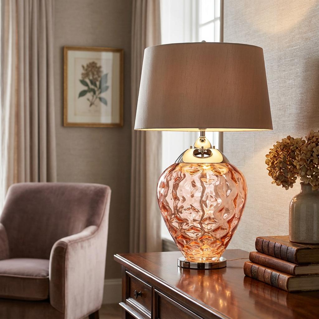 Samara salmon pink glass table lamp with a taupe drum shade on a wooden desk in a cozy living room.