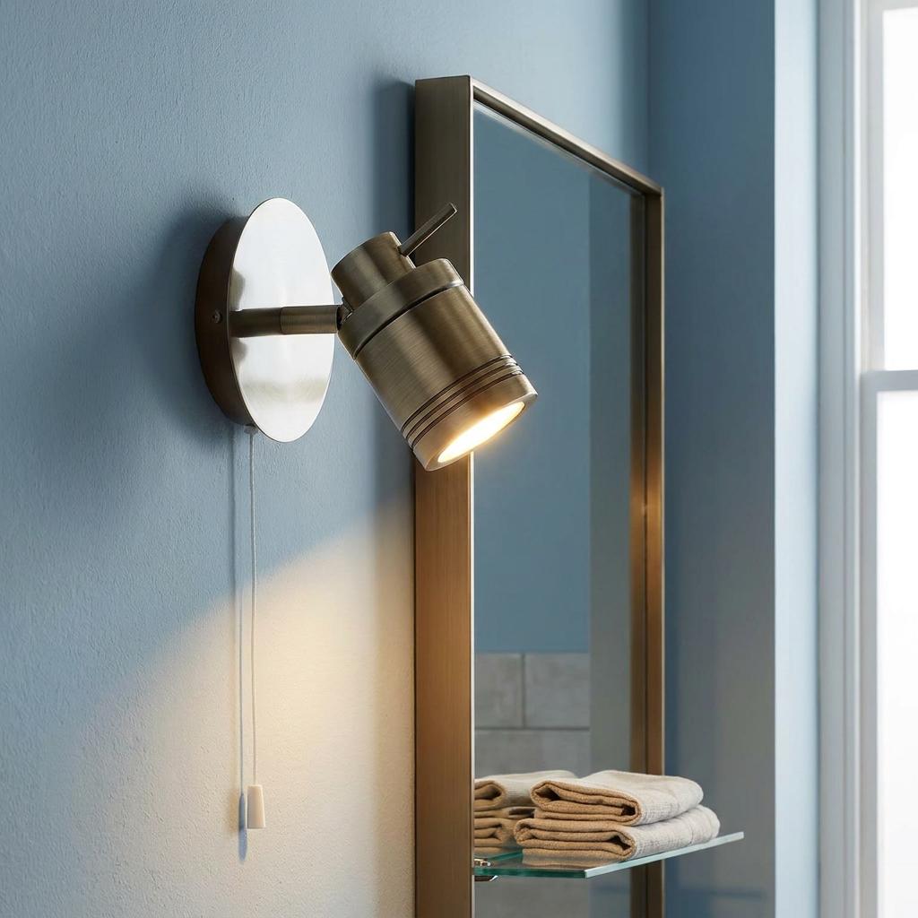 An illuminated brass bathroom spotlight with a white pull cord mounted next to a framed mirror on a light blue wall.