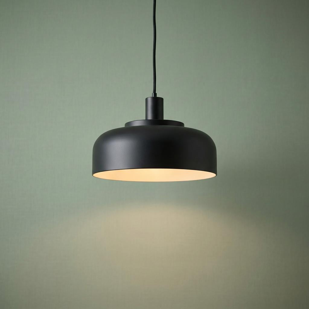 Matt black pendant light with a rounded dome shade hanging against a textured sage green background.