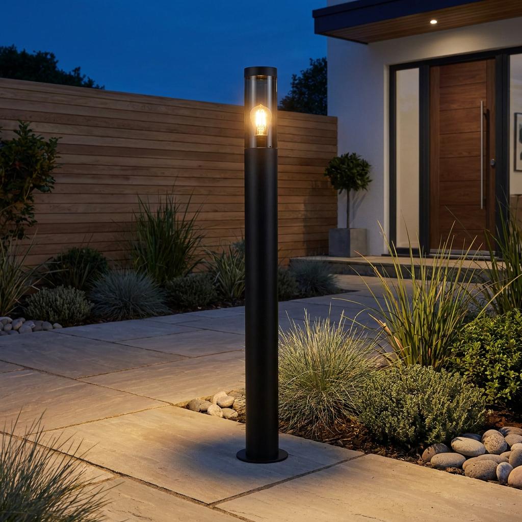 A black cylindrical outdoor bollard light illuminating a paved garden walkway at night.