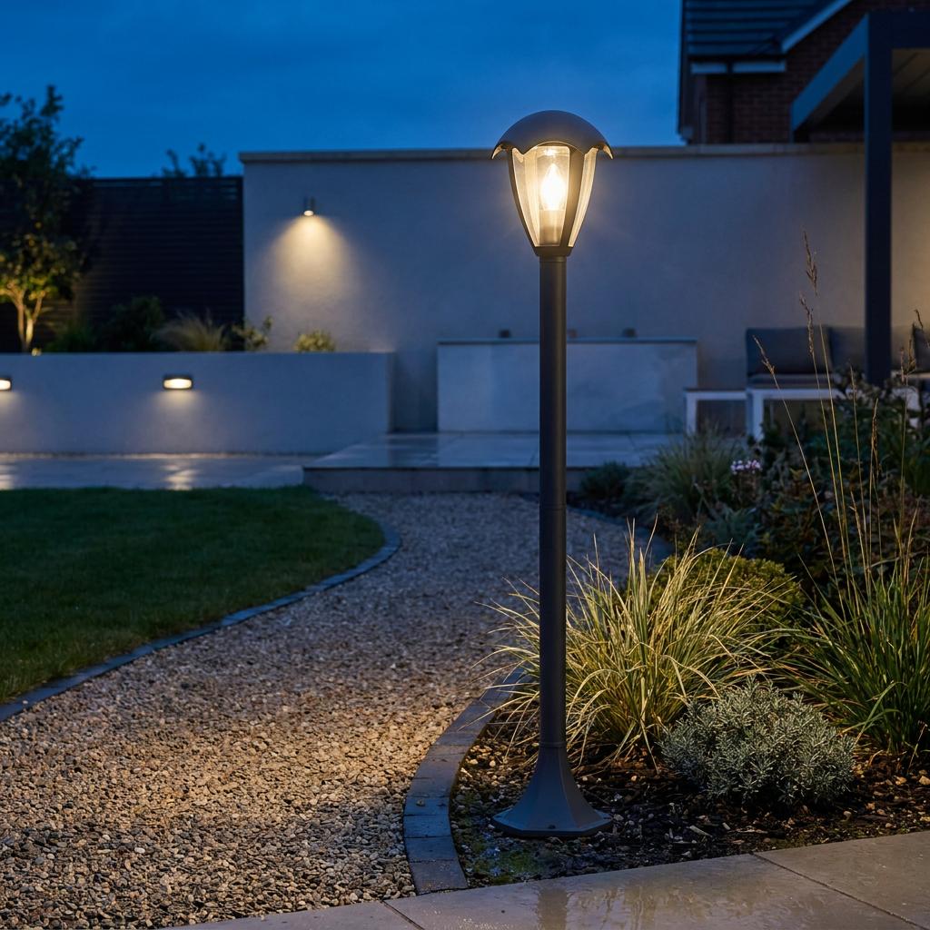 Modern dark grey lantern-style bollard light glowing beside a gravel garden path at twilight.