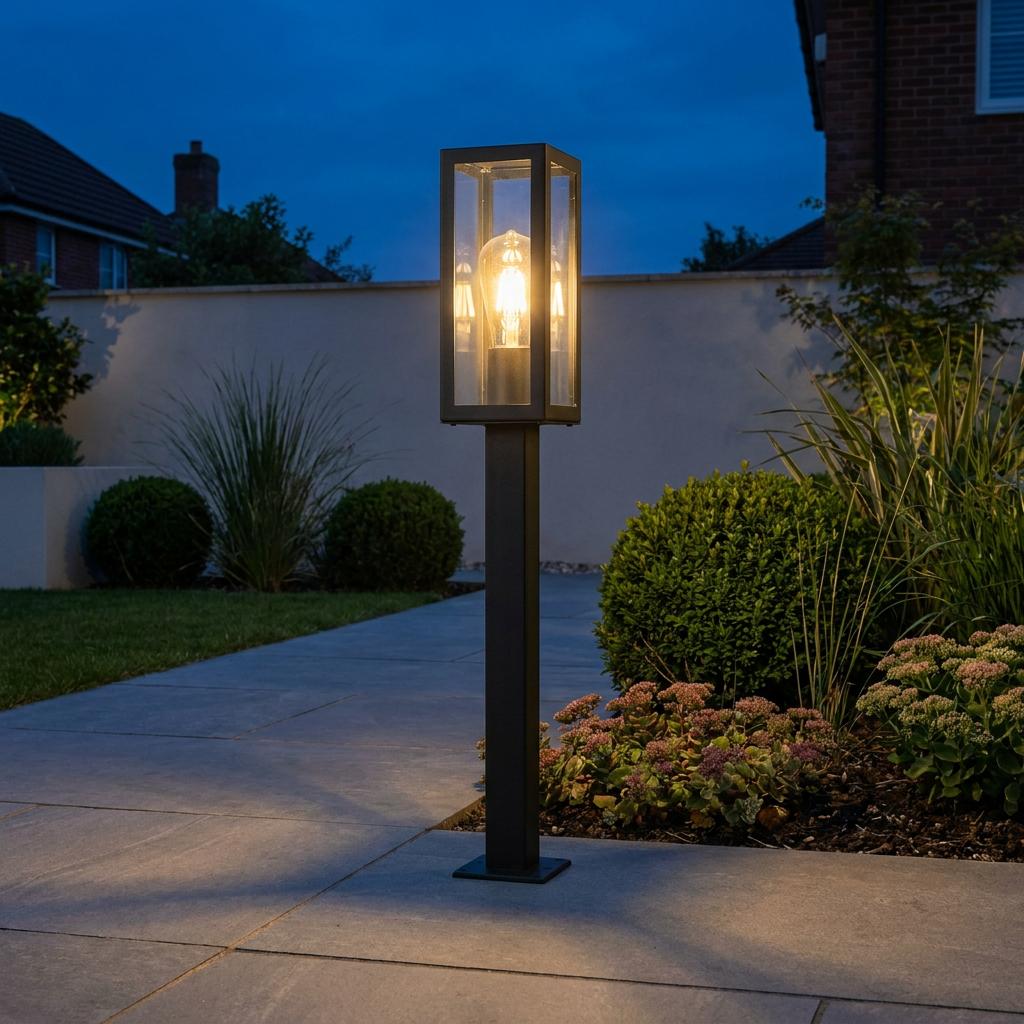 Modern black bollard light with a glowing filament bulb illuminating a garden pathway at dusk.