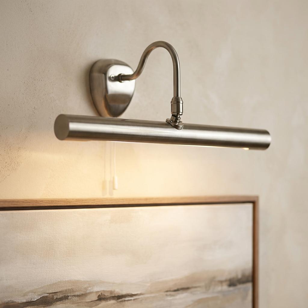 Satin nickel adjustable picture light mounted on a wall, casting a warm glow over a framed landscape painting.