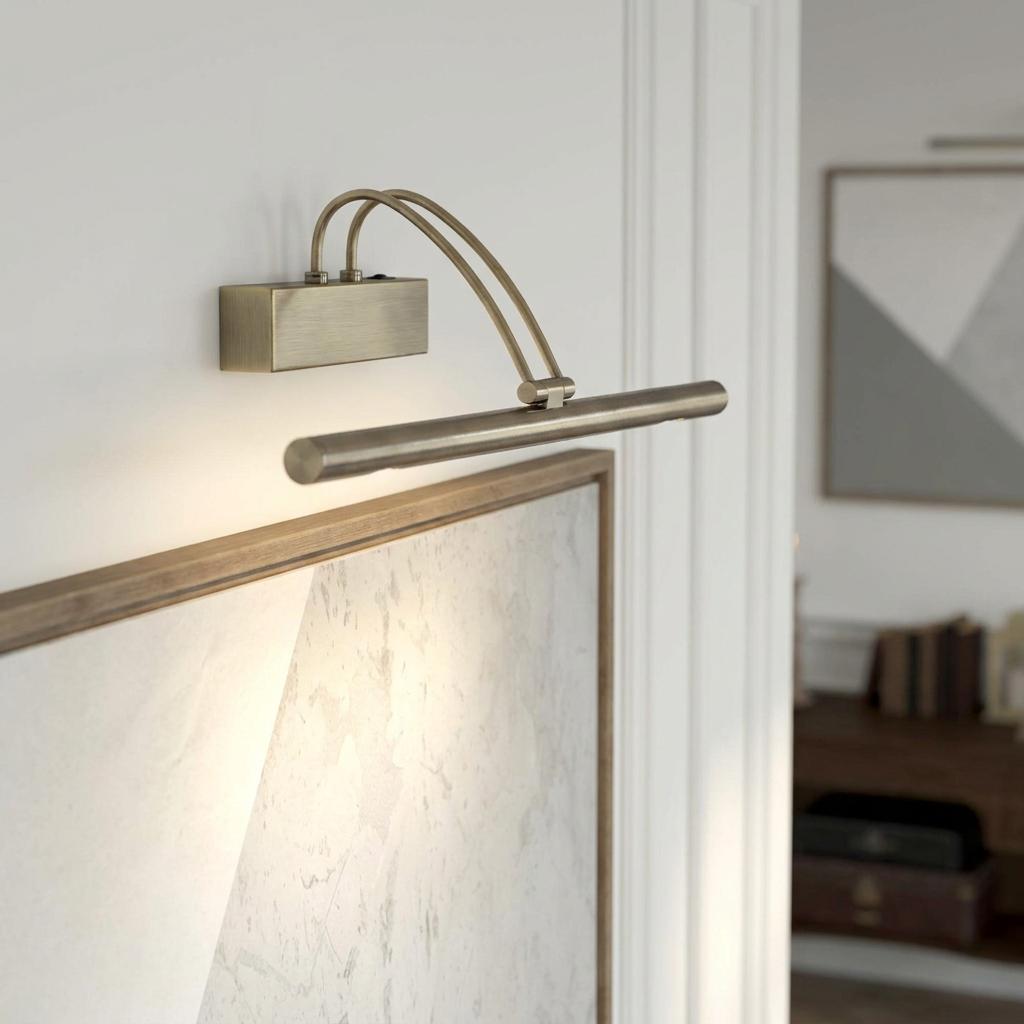 Slimline antique brass picture light with adjustable twin arms mounted on a white wall above a framed artwork.